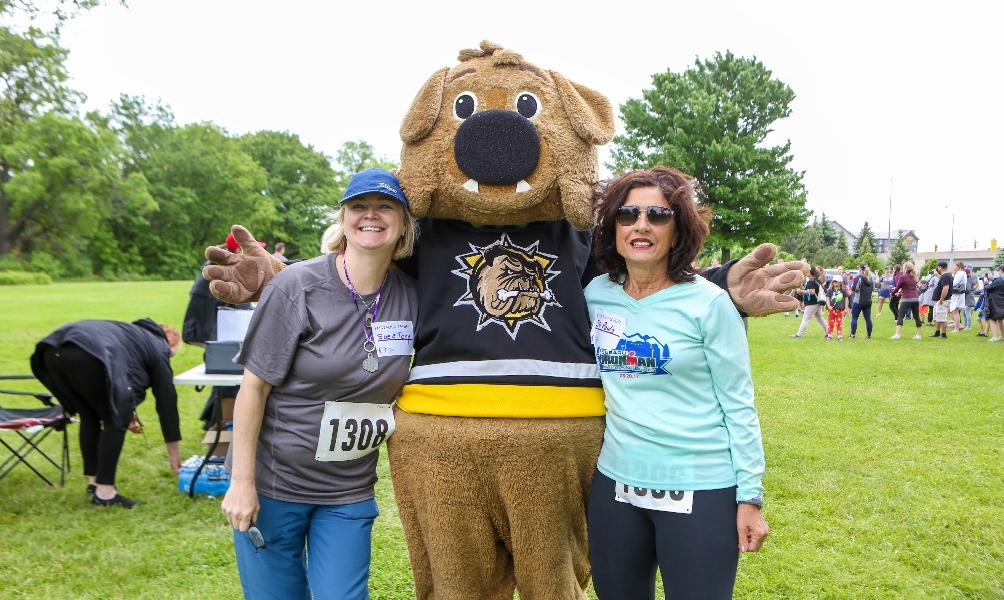 Two women wearing runner numbers on their clothes pose with the Hamilton Bulldogs mascot at a fundraising event
